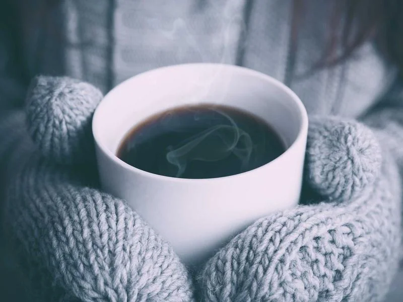 A steaming cup of tea held in knitted mittens